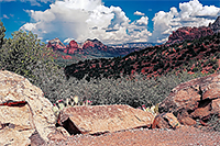 White fluffy clouds against a blue sky with red mountains and light colored rocks in the foreground