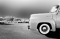 A front end side view of a 1950s Ford pickup on asphalt with sun gleaming